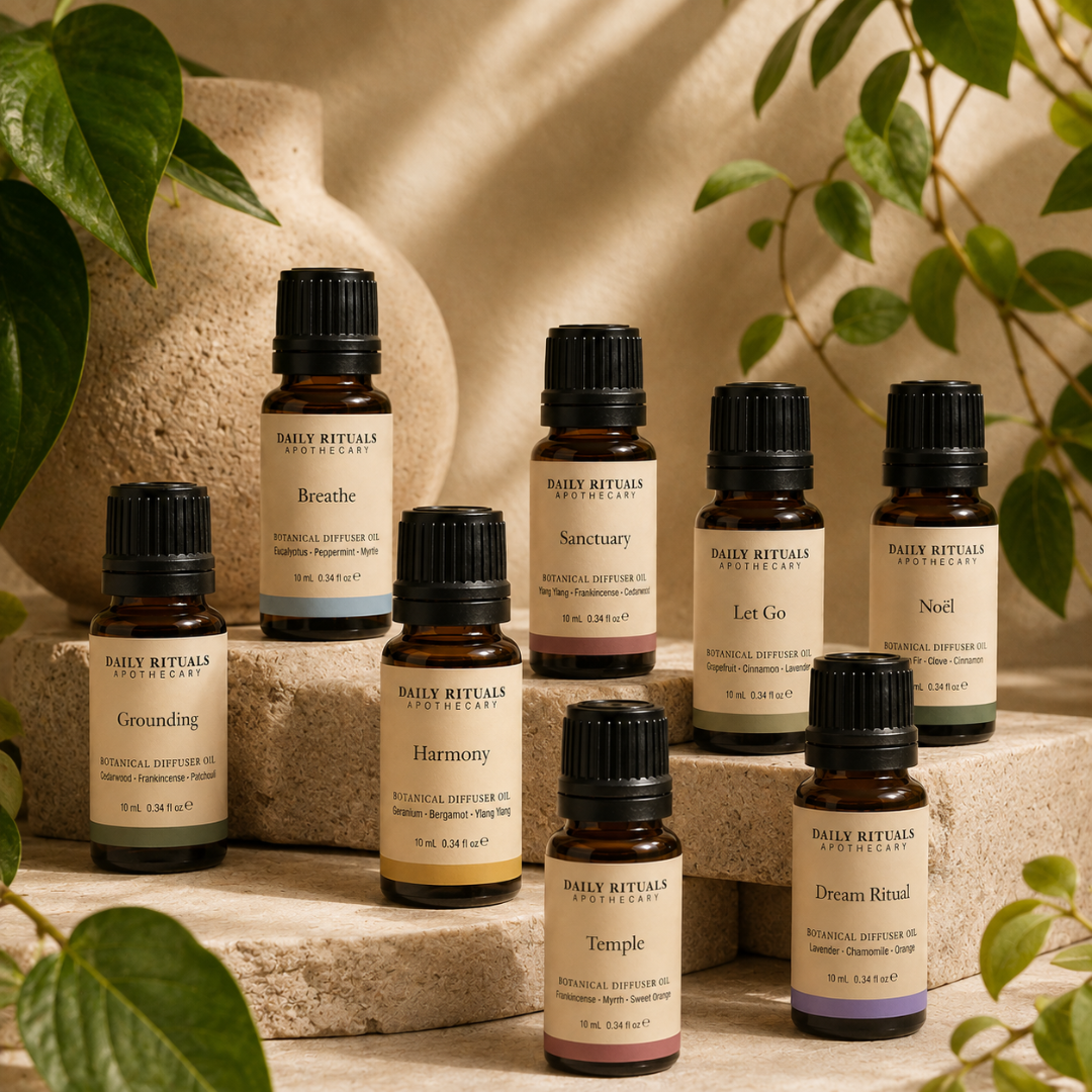 A set of essential oil bottles from Daily Rituals Apothecary arranged on stone steps with green leaves and a textured stone wall in the background. The bottles are amber glass with black caps and cream labels featuring different colored accents.