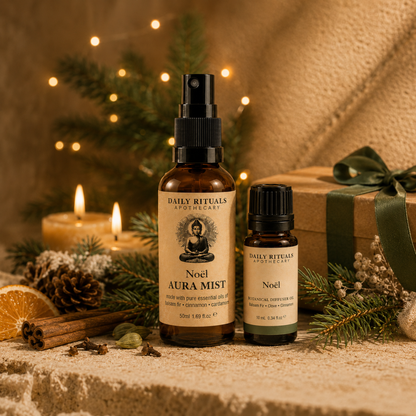 Two amber glass bottles from Daily Rituals Apothecary labeled 'Noel Aura Mist' and 'Noel' are styled on a stone surface with holiday greenery, candles, cinnamon sticks, and a gift box in the background.